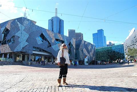 Woman standing beside the geometric facade of Federation Square, Melbourne, under a blue sky