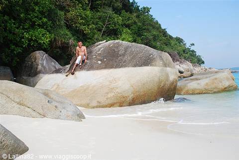 Young man sitting on huge granite boulders between white sand and turquoise sea