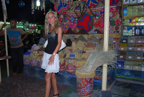 Blonde woman in white skirt in front of spice stall with decorated baskets at night