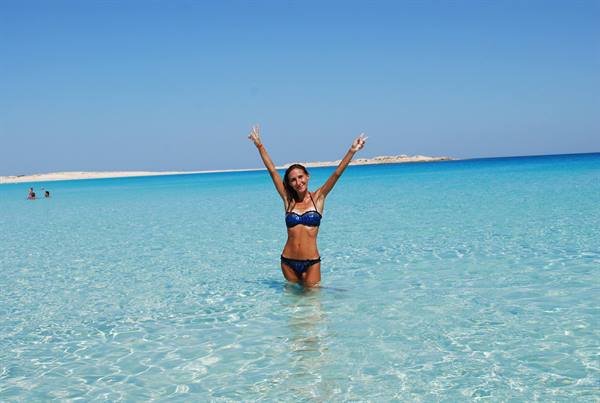 Woman in bikini raising arms in turquoise water, white beach behind