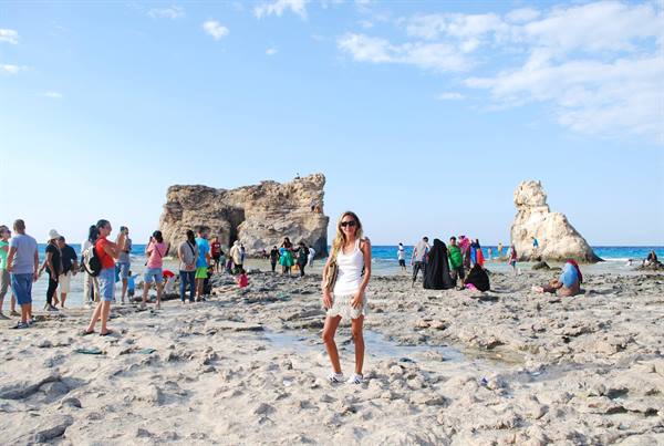 Woman posing on rocky beach with limestone formations and swimmers under blue sky