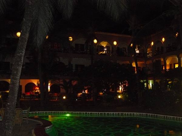 Green-lit pool at night among palm trees and arched hotel balconies