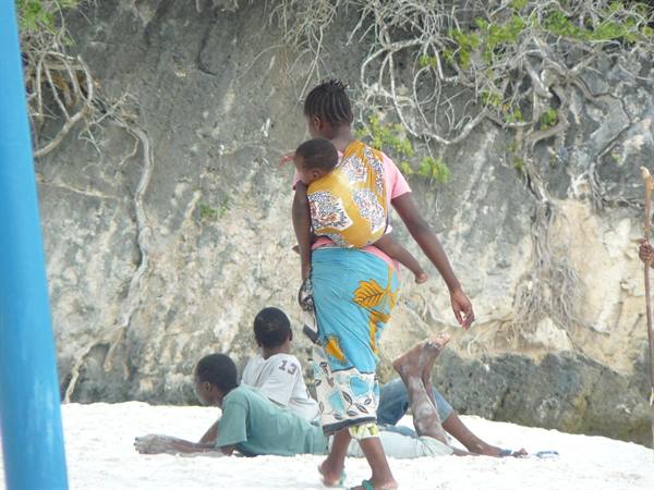 Woman carrying a child on her back walks on white sand past resting boys