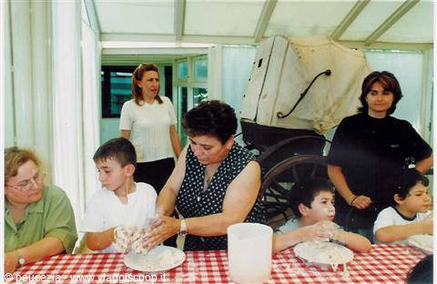 Children kneading dough at a table, old covered cart behind in glass room