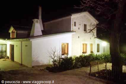 White stone farmhouse lit at night, glowing windows behind bushes