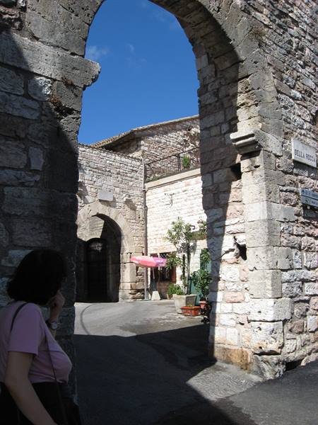 Medieval limestone arch, people passing by, sharp shadows under blue sky