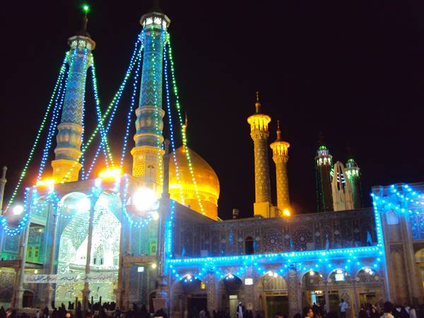 Illuminated shrine at night: golden dome, blue-tiled minarets, string lights, crowd