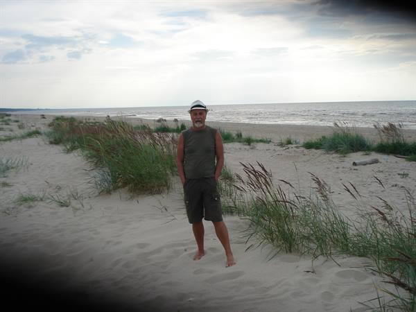 Barefoot man in hat among sandy dunes and grass, facing the sea