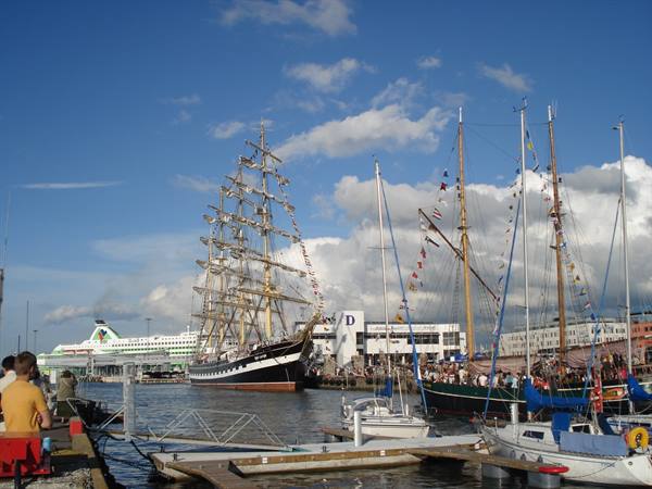 Three-masted tall ship docked among sailboats, crowd on pier under clouds