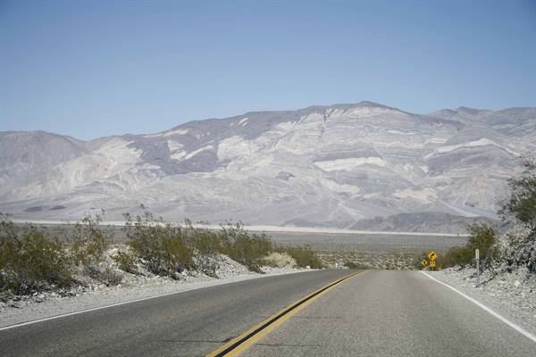 Paved road through desert shrubs toward pale rocky mountains, blue sky