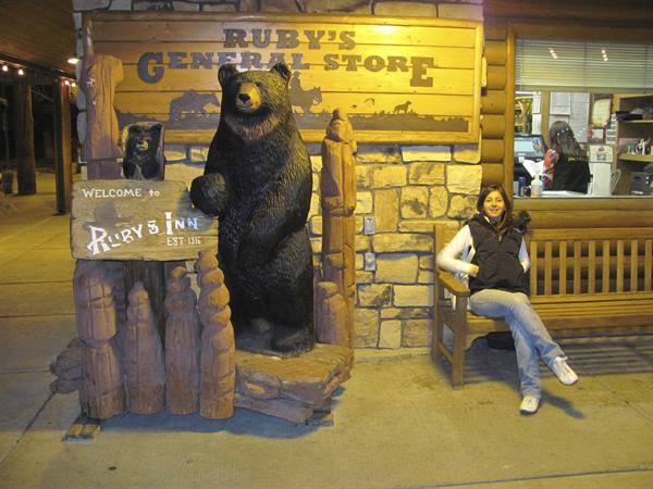 Woman sits on wooden bench beside carved bear at Ruby's General Store