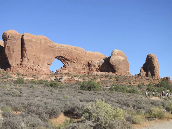 Visitors walk among desert shrubs beneath the red North Window rock arch, Arches