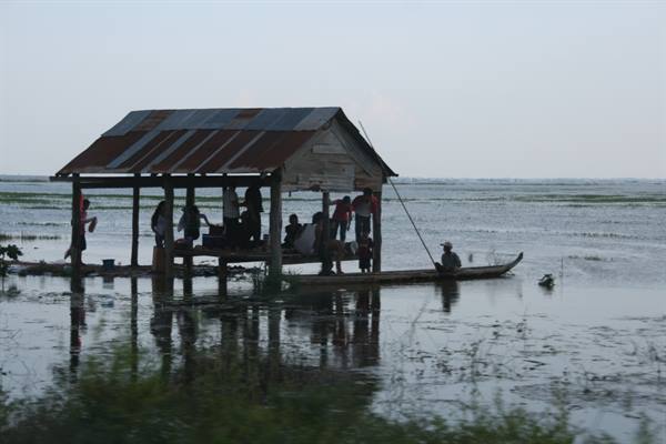 Stilt wooden hut on calm water, people under corrugated roof, fisherman on boat