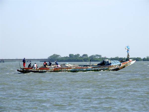 Wooden pirogue loaded with men and goods glides on brown water, bridge and trees behind