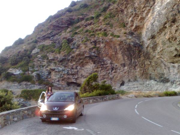 Woman stepping out of a car on a coastal road, steep rocky cliff behind