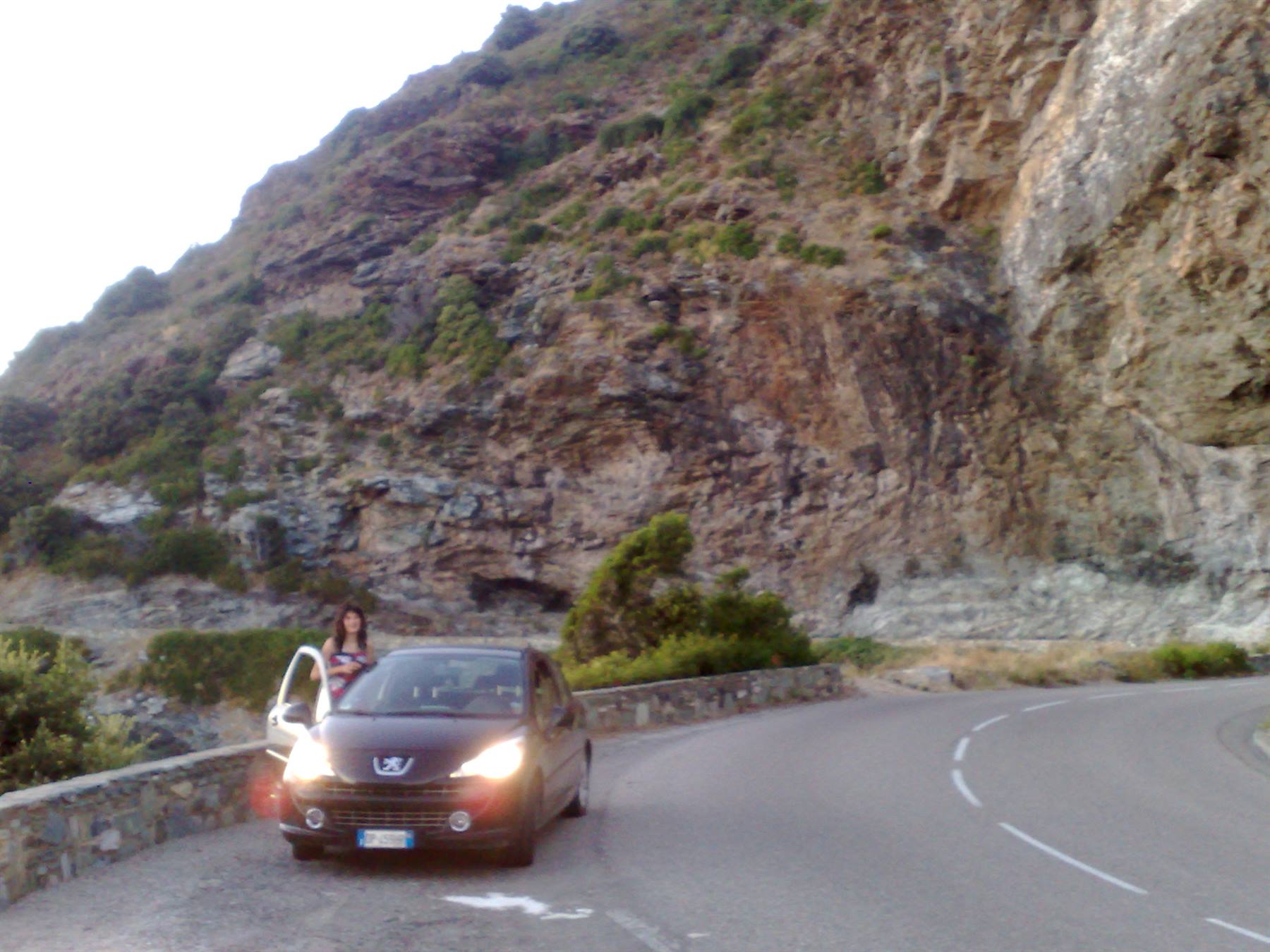 Woman stepping out of a car on a coastal road, steep rocky cliff behind