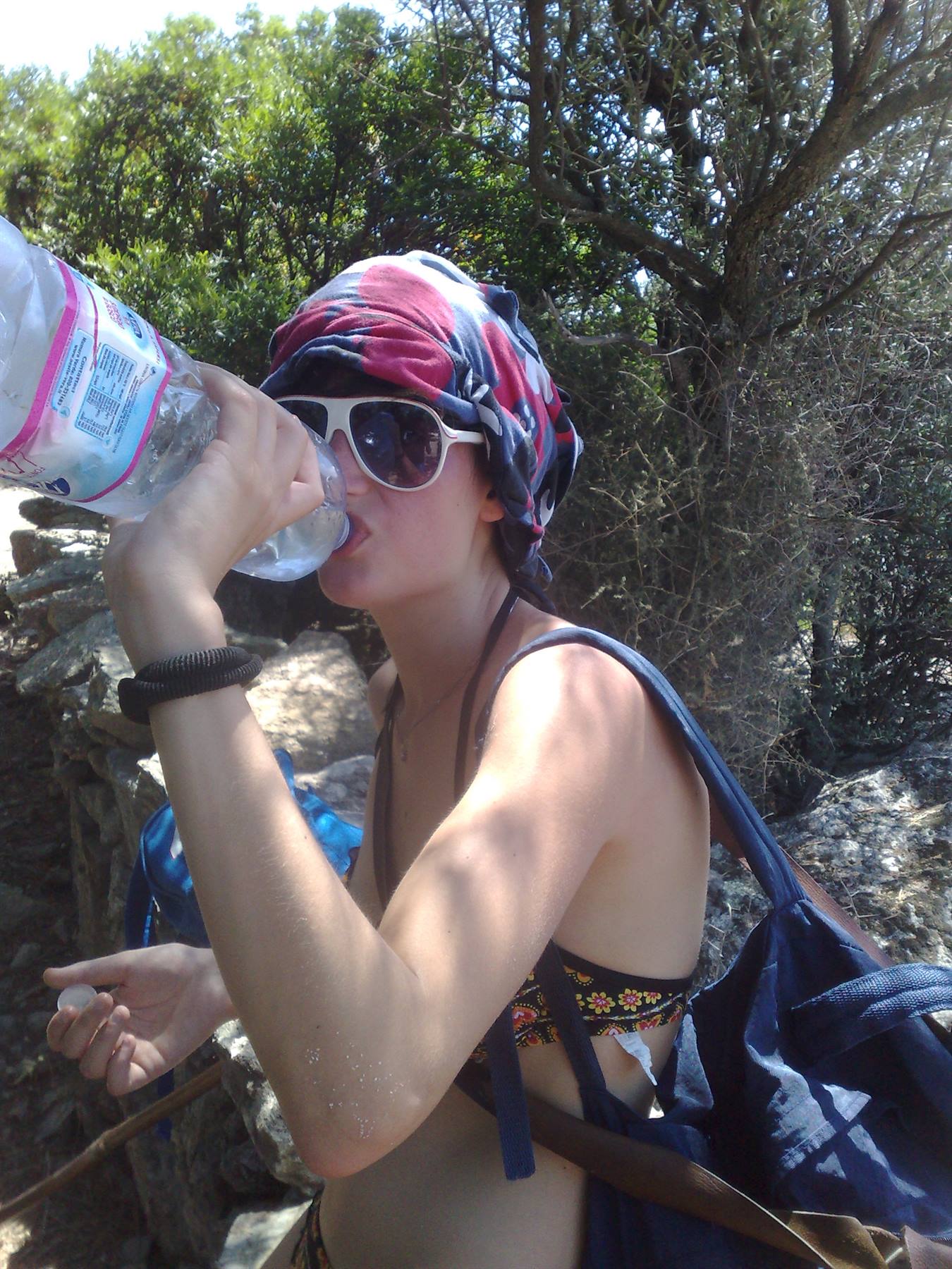 Girl in headscarf and sunglasses drinks water on a sunny rocky trail