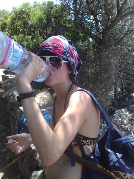 Girl in headscarf and sunglasses drinking water among Mediterranean shrubs and stone walls