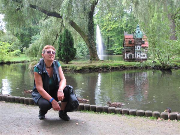 Woman crouching by a pond with ducks, fountain and small historic house