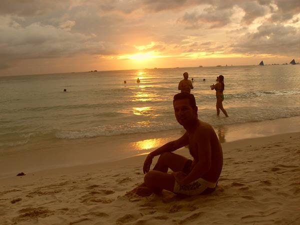 Man sitting on the sand at sunset, swimmers and sailboats on the sea