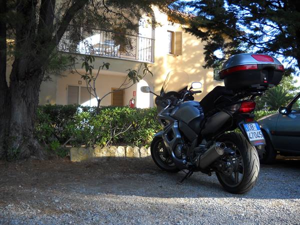 Grey touring motorcycle with top box parked on gravel by a yellow house