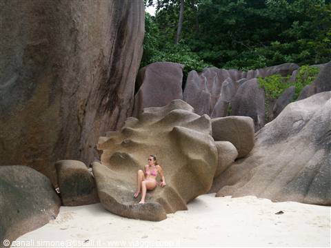 Woman in bikini sitting on sculpted granite boulder, white sand beach, tropical vegetation behind