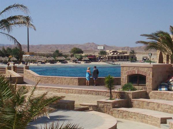 Couple on stone wall by turquoise lagoon, palm trees and beach umbrellas, Red Sea