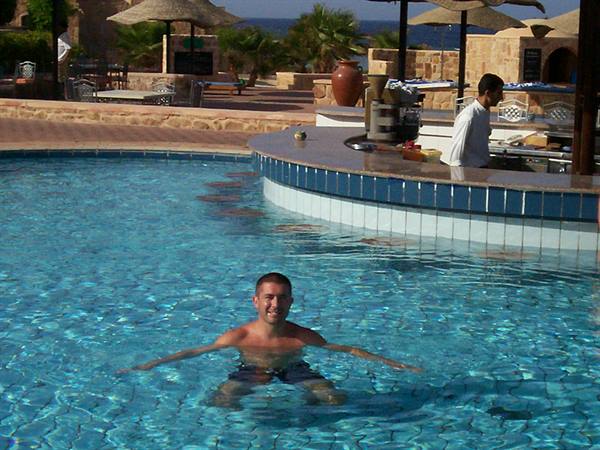 Man in swim trunks swims in pool, poolside bar and straw umbrellas behind