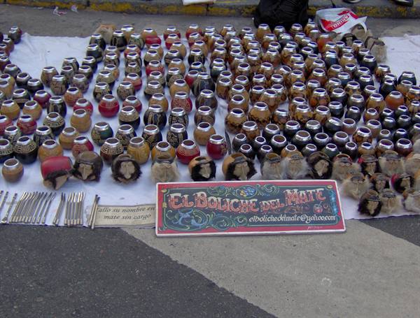 Street stall displaying many painted mate gourds on cloths, sign El Boliche del Mate
