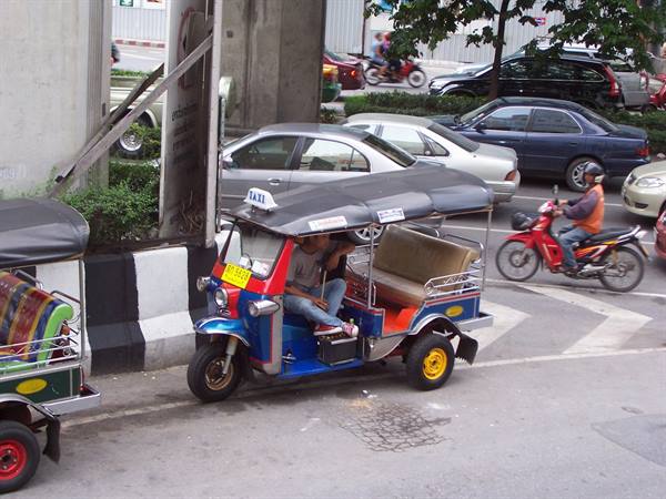 Blue and red tuk-tuk parked at the curb, motorcyclist passing between cars