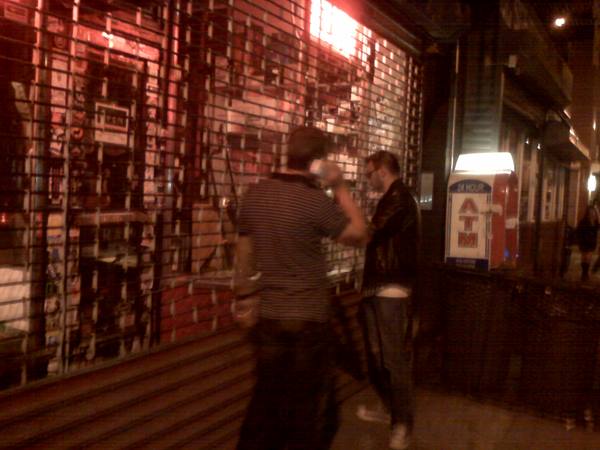 Two men drinking at night in front of a shuttered storefront, next to an ATM