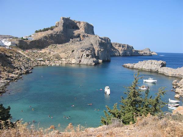 Swimmers in Lindos turquoise bay, rocky cliffs and white boats under sun