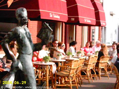 Bronze statue holding a book in front of a café terrace with wicker chairs