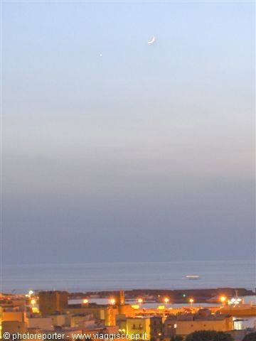 Crescent moon above the lit town and calm sea at dawn
