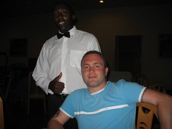 Waiter in white shirt and bow tie smiles beside seated tourist, dim indoor light