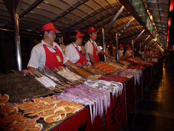 Night food market: vendors in red aprons serve starfish and squid skewers