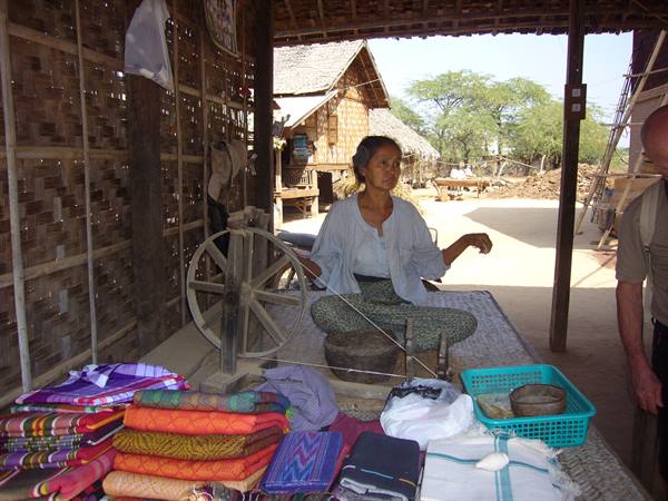 Woman spinning silk at wooden wheel, colorful fabrics for sale in the village