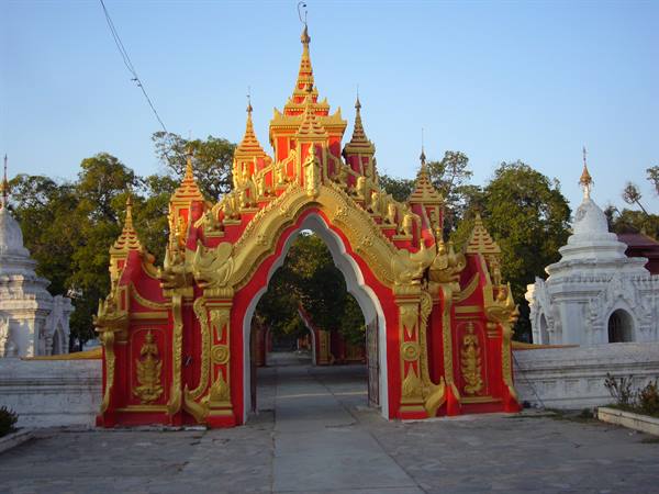 Red and gold temple gateway with ornate arch, white stupas beside, green trees
