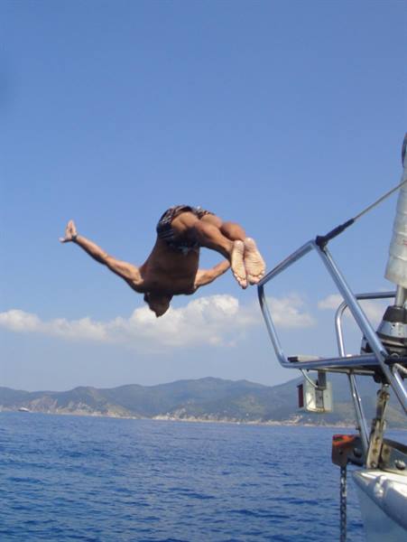 Man diving mid-air off steel yacht railing, blue sky, coast and mountains beyond