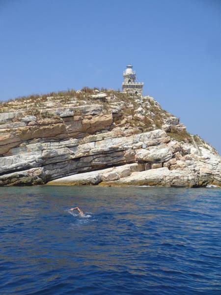 Swimmer in blue water below the rocky cliff and Tinos lighthouse