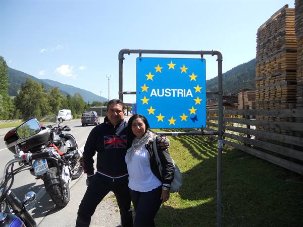 Motorcycle couple in front of blue AUSTRIA sign, stacked timber and mountains
