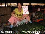 Smiling woman sells vegetables at a market stall beside bananas and greens