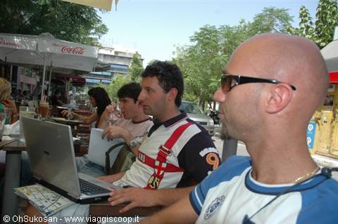 Two men at an outdoor café looking at a laptop under an umbrella