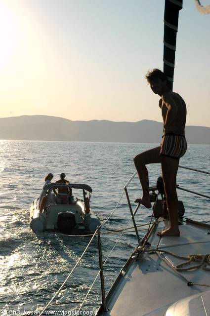 Boy in swimsuit standing on sailboat deck, dinghy approaching at sunset