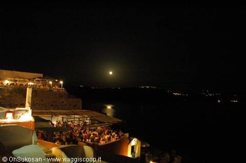 Full moon over Santorini caldera, crowded terrace lit up at night
