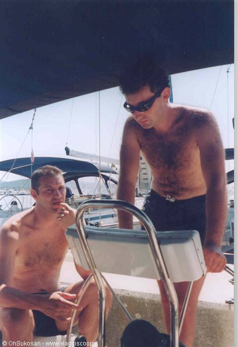 Two shirtless men aboard a sailboat in the marina, cigarette and stainless wheel