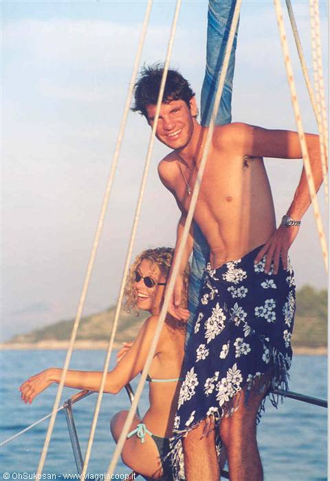 Smiling couple in swimwear aboard a sailboat among rigging, green coastline behind