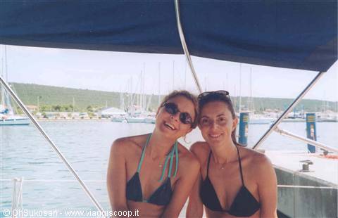 Two girls in bikinis under the blue canopy of a sailboat, marina behind