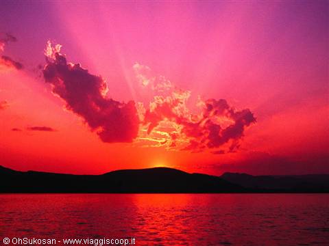 Red sun rising behind dark hills, pink clouds glowing over the sea