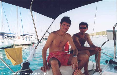 Two young men aboard a sailboat at a marina, steering wheel visible